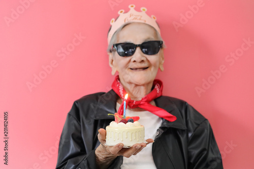 Senior Asian woman celebrating a birthday while holding a small cake with a lit candle against a vibrant pink background.
