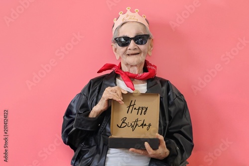 Senior Asian woman celebrating a birthday while holding pizza box with wording Happy Birthday against a vibrant pink background.