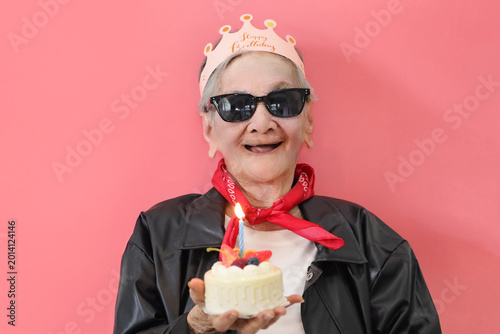Senior Asian woman celebrating a birthday while holding a small cake with a lit candle against a vibrant pink background.