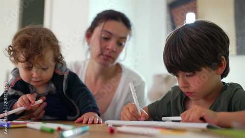 Foto Baby explores markers beside older brother drawing at table while mother remains