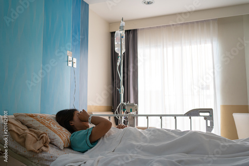Fotografie A stressed Asian female patient lying in a hospital bed, covering her eyes in pain or grief