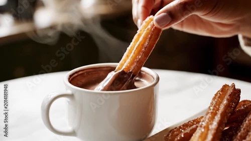 A person's hand reaches for a plate of golden fried churros dusted with sugar, served next to a steaming white cup of hot chocolate with a spoon on a white table in a cafe setting.