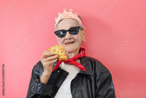 Fashionable senior woman enjoying a slice of pizza during a birthday celebration against a bright pink background.