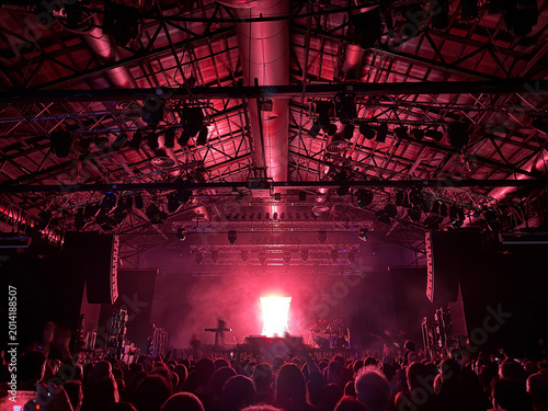 High-energy rock concert crowd gathered in a large, packed music venue. A band performs on stage under dramatic, colorful lighting, with hundreds of people cheering and raising their hands
