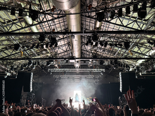 High-energy rock concert crowd gathered in a large, packed music venue. A band performs on stage under dramatic, colorful lighting, with hundreds of people cheering and raising their hands