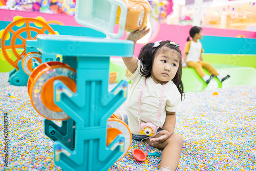 Schilderij op canvas Cute little Asian girl playing in a colorful ball pit at an indoor playground,A lovely young Asian girl with pigtails enjoying her time sitting among thousands of colorful small plastic balls