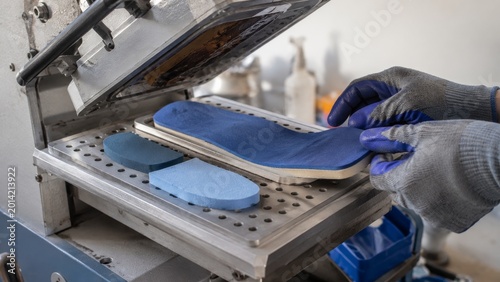Gloved hands position a blue orthopedic insole on a heated press machine with foam inserts on a perforated metal platform in a workshop