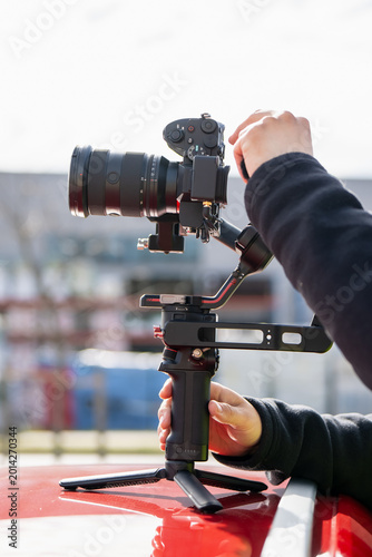 Professional filmmaker balancing a mirrorless camera on a handheld three-axis gimbal stabilizer placed on a red car roof for smooth cinematic video production in urban setting