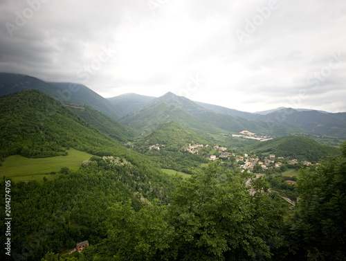 Tableau sur toile Panorama montano con alberi in primo piano, valle verde e piccolo centro abitato
