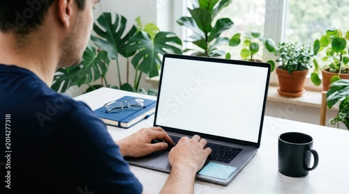Over the shoulder view of a man typing on a laptop with a white blank screen at a bright desk with green plants in the background.