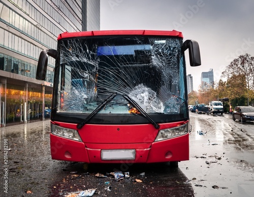 a red city bus is displayed with a severely shattered windshield surrounded by debris indicating it has been involved in a serious accident on a rainy urban street