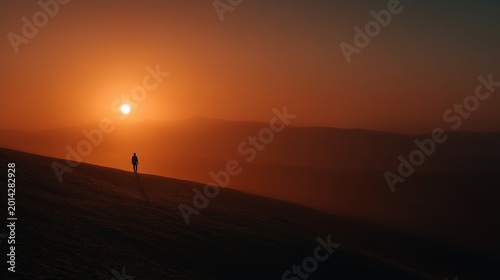 A solitary person walks on a vast dune at sunset, casting a long shadow against a dramatic orange sky.