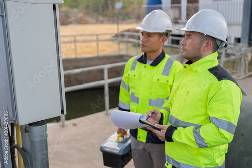 Two Asian engineers inspecting electrical control cabinet at irrigation waste trap station. Professional technicians checking water management system and monitoring hydraulic equipment outdoors.