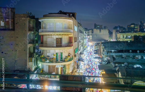 A high angle view of a bustling night market in Cairo, Egypt, captured from a passing bus on an elevated road, showing illuminated buildings and crowds.