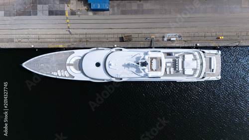 Top down aerial view of a large white luxury yacht docked at a harbor quay. Modern maritime scene with dark water, marina infrastructure and elegant vessel design seen from above.