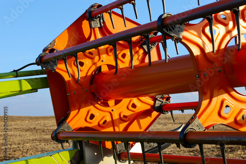 A close-up of a bright orange reel on a modern grain harvester focuses on its intricate design and metal components. This agricultural equipment sits in an empty field under a clear sky, ready for wor