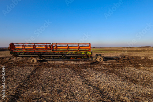​A powerful combine harvester header sits on a trailer in the middle of a wide mown field under a clear blue sky. Agricultural machinery is ready for transport after the grain harvest is complete.