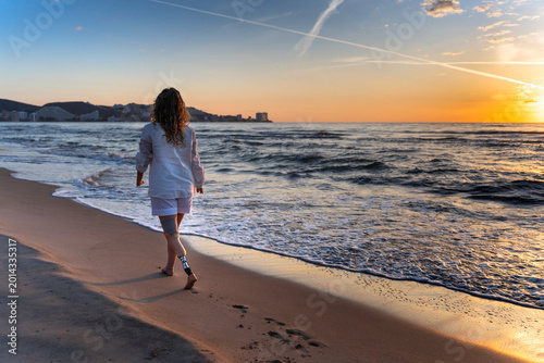 Woman walking with prosthetic leg on beach at sunset, showing inclusion and freedom