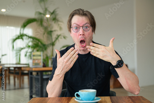 Authentic portrait of a shocked Scandinavian man showing disbelief while sitting in a cafe with glasses.