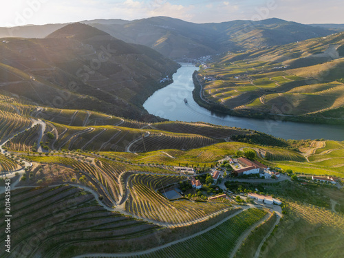 Tour boat passes through the Douro River below terraced vineyards and estates in Portugal's Douro Valley at sunset.