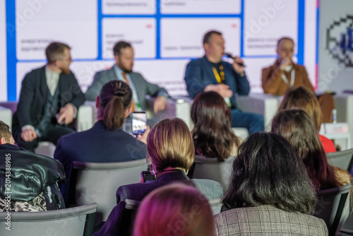 Audience watches panel discussion at business conference, attendee records speakers with smartphone, networking vibe and focused engagement.