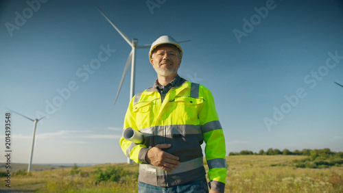 Caucasian male engineer wearing helmet and reflective jacket holding rolled blueprint standing near wind turbines. Worker looking directly at camera in open field on sunny day.