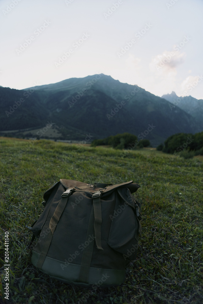 Fototapeta premium Backpack resting on a grassy meadow with distant mountains and clear sky. Focus on outdoor gear, nature exploration, and rugged solitude in a calm landscape.