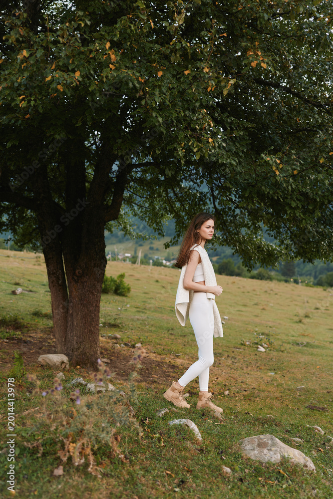 Naklejka premium Relaxed woman strolling through a sunlit countryside, dressed in white, enjoying fresh air and nature, a casual lifestyle moment by a large tree.