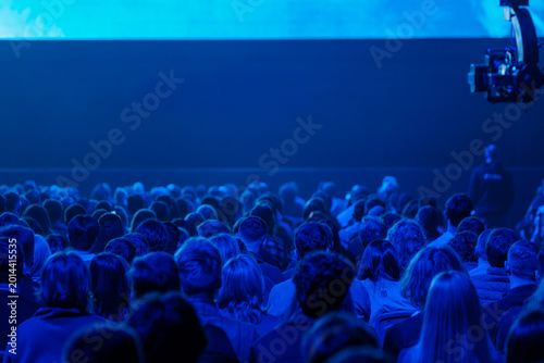 Large audience seated in blue lit conference hall, watching presentation on stage. Moody lighting, crowded rows, live event atmosphere and focused attention.