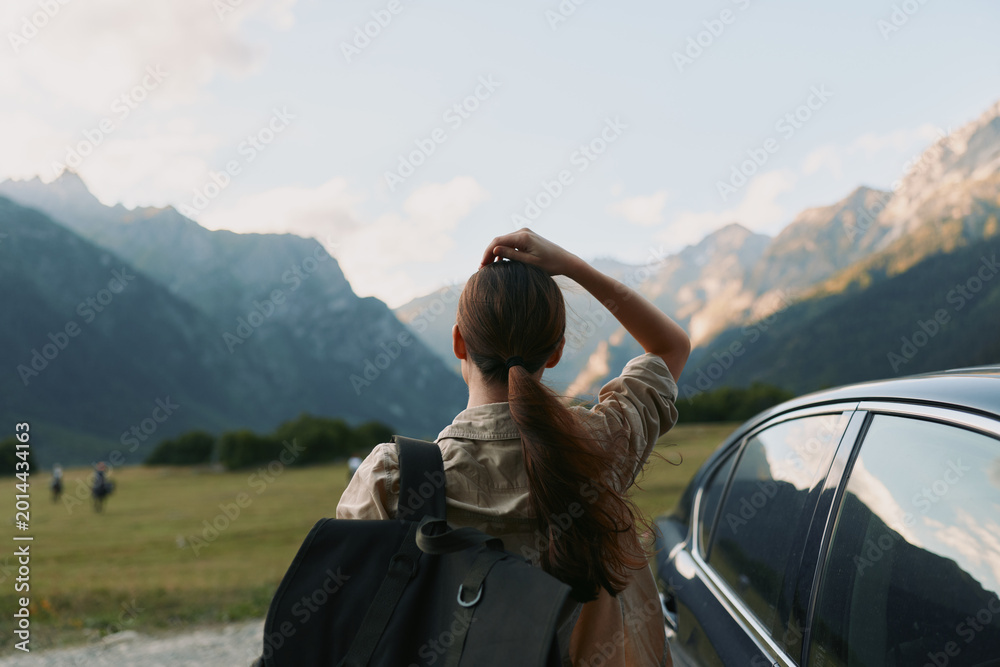 Naklejka premium Woman with backpack standing beside a parked car, gazing at distant mountains, enjoying outdoors lifestyle, ready for travel and adventure in a scenic valley landscape.