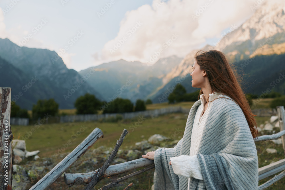 Fototapeta premium Young woman with long hair gazes at alpine mountains, wrapped in a cozy shawl, standing beside a weathered wooden fence, enjoying a calm lifestyle moment in nature