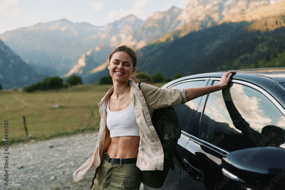 Naklejka premium Woman enjoying outdoor lifestyle beside a car in mountain landscape, smiling confidently, wearing casual hiking attire, sunlit scenery and sense of adventure