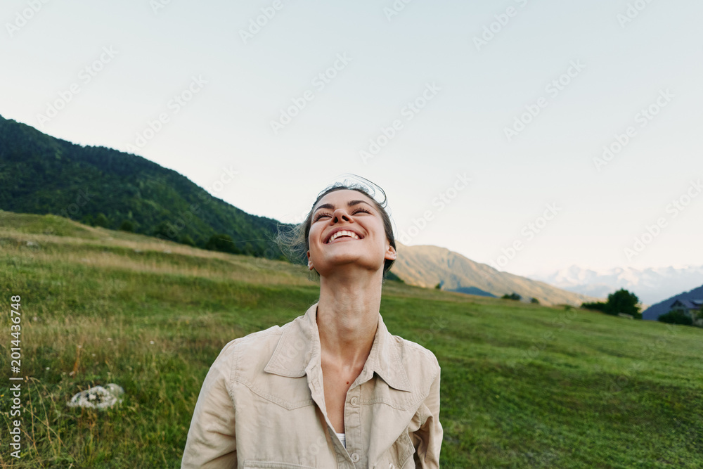 Fototapeta premium Cheerful woman outdoors, smiling up at the sky in a sunlit meadow, relaxed posture, natural colors, wide landscape, mountains in distance, lifestyle moment