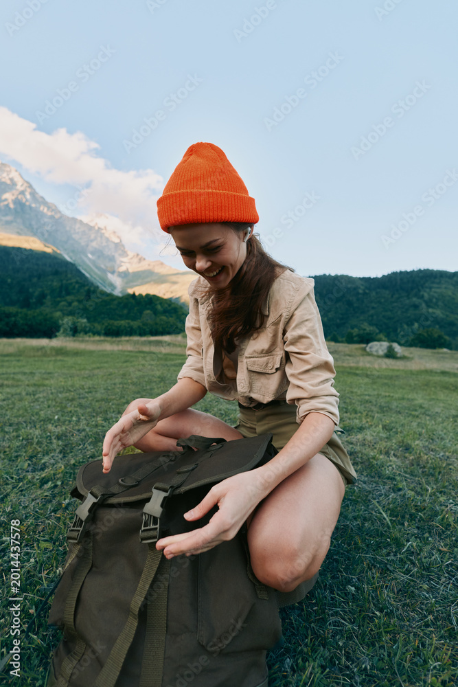 Fototapeta premium Woman in an orange beanie sits on grass beside a backpack in a mountain meadow, enjoying outdoor life and natural scenery and sunlight warms her face under a clear blue sky