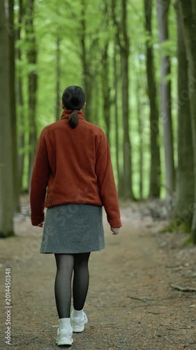 Back view of a young woman in a bright orange sweater and skirt walking along a forest path and stopping to breathe fresh air in a peaceful natural environment.