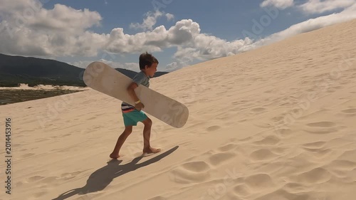 Young boy walks up sand dune mountain with sand board to slide down - vacation fun