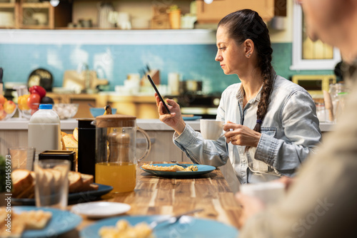 Fotografía Mom engrossed in her smartphone screen due to social media addiction, being detached and ignoring her family at breakfast