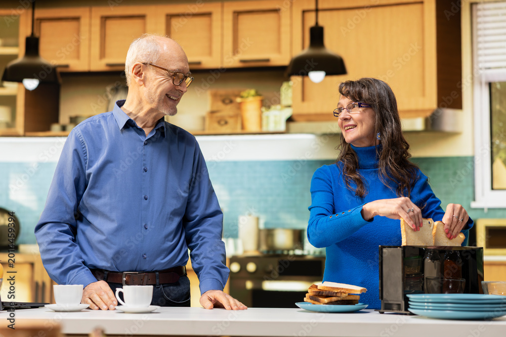 Fototapeta premium Senior couple preparing breakfast together in a modern kitchen, cozy morning routine. Husband and wife sharing a candid moment at kitchen counter, serving healthy food and warm coffee.