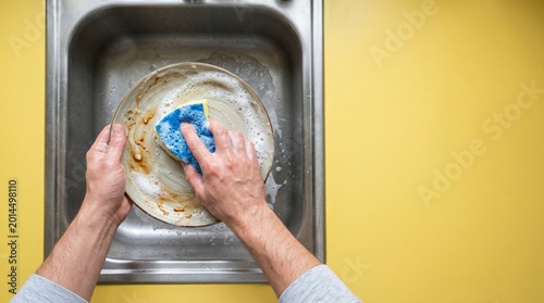 Top view of hands washing a dirty ceramic plate with a sponge on a yellow background with copy space
