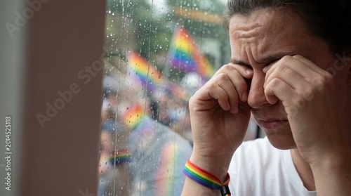 Young woman crying while looking through rainy window at pride parade celebration outside