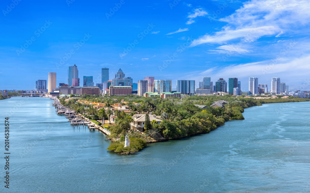 Fototapeta premium Aerial view of Tampa, Florida, featuring a lush waterfront peninsula with luxury homes, private docks, a small lighthouse, and the downtown city skyline in the background