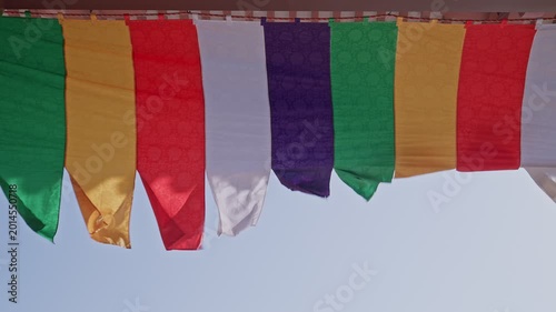 Vivid Buddhist ceremonial banners in gold, red, white, purple, and green sway gently in the wind beneath temple eaves against a pale blue sky, their fabric edges rippling with movement.