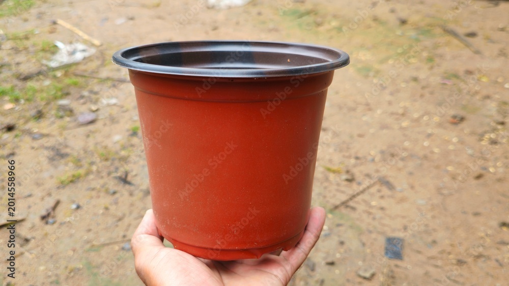 Naklejka premium Hand Holding Empty Brown Plastic Plant Pot on Outdoor Background, Gardening Container. Hand holding empty red plastic flower pot on ground background.