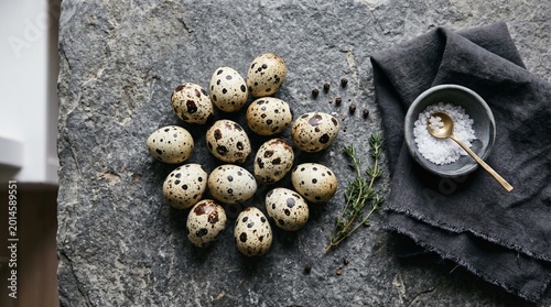 Flat lay of fresh speckled quail eggs on rustic stone background with sea salt and herbs.