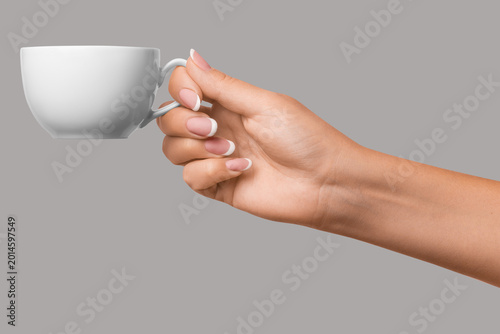 Fotografi Hand with manicured nails holding white teacup on gray background, elegant and m