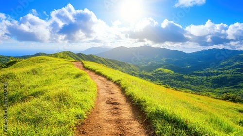 A sunlit mountain hiking trail winds through lush green hills towards a hazy horizon, offering breathtaking scenic vistas.