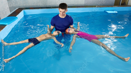 Fényképezés Little children learning how to swim in pool with teacher, laying on he back