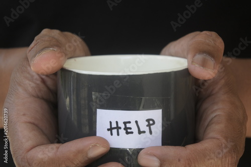 Fotografi Close up of wrinkled hands holding a gray mug with a HELP sign taped to it, representing poverty, hunger, and the need for social assistance