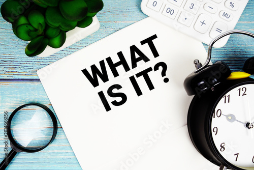 What Is It? written on paper placed on a blue wooden surface, alongside a magnifying glass, a clock, a calculator, and a small plant