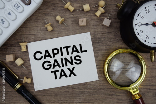 Capital Gains Tax text written on notepad with pen, alarm clock, magnifying glass and calculator on wooden background